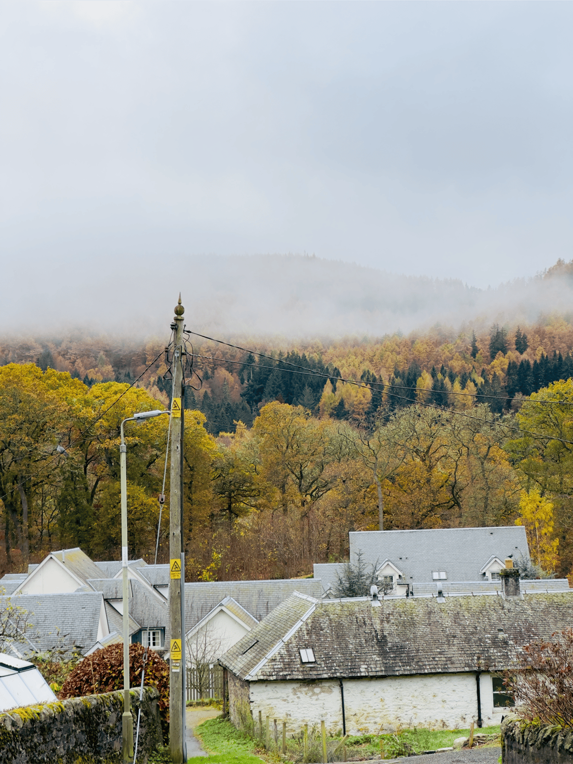 A serene Pitlochry, Scotland during November autumn, with mist covering colorful forested hills and rooftops of stone cottages in the foreground.