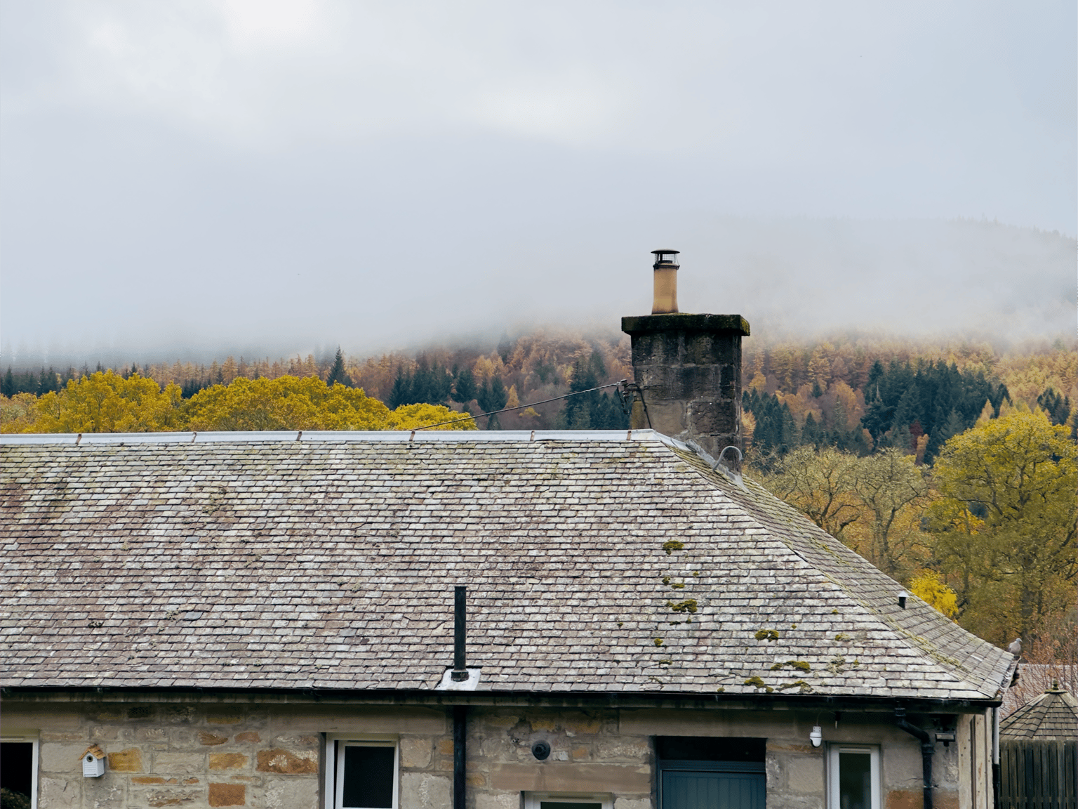 A serene Pitlochry, Scotland during November autumn, with mist covering colorful forested hills and rooftops of a stone cottage in the foreground.