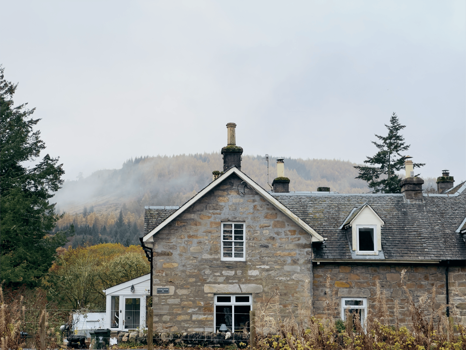 A serene Pitlochry, Scotland during November autumn, with mist covering colorful forested hills and a stone cottage in the foreground.