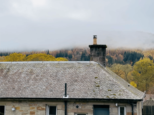 A serene Pitlochry, Scotland during November autumn, with mist covering colorful forested hills and rooftops of a stone cottage in the foreground.